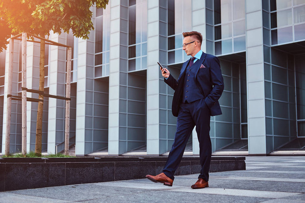 Портрет уверенного стильного бизнесмена, одетого в элегантный костюм с помощью смартфона, стоящего на открытом воздухе на фоне небоскреба
. - Фото, изображение