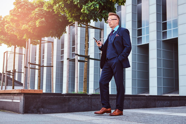 Portrait d'un homme d'affaires élégant et confiant vêtu d'un costume élégant tient un smartphone et regarde loin tout en se tenant debout à l'extérieur sur un fond de gratte-ciel
. - Photo, image