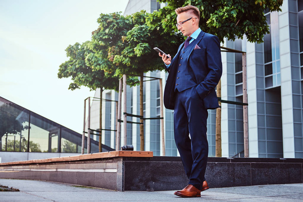 Retrato de un hombre de negocios con estilo y confianza vestido con un traje elegante usando un teléfono inteligente mientras está parado al aire libre contra el fondo del rascacielos
. - Foto, imagen