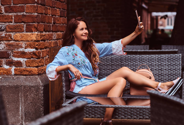 Чуттєва жінка з довгим коричневим волоссям в модному одязі робить селфі сидячи в кафе
. - Фото, зображення