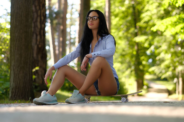 Gorgeous sensual brunette girl in glasses wearing a shirt and shorts sitting on a skateboard in the park. - Photo, Image