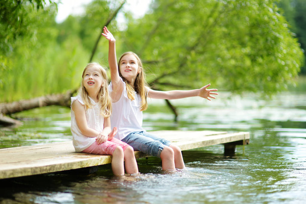 Two cute little girls sitting on a wooden platform by the river or lake dipping their feet in the water on warm summer day. Family activities in summer. - Foto, Imagem