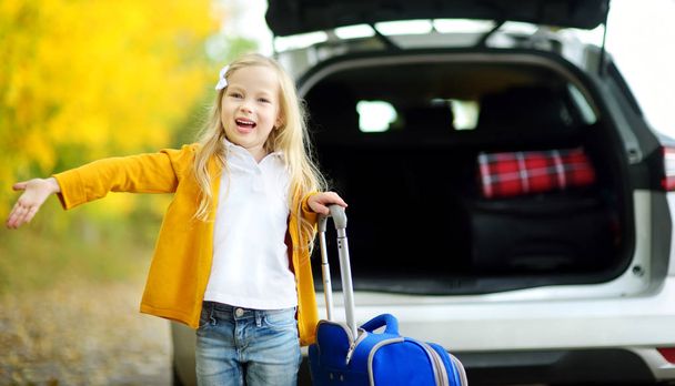 Adorable girl with a suitcase ready to go on vacations with her parents. Child looking forward for a road trip or travel. Autumn break at school. Traveling by car with kids. - Фото, изображение