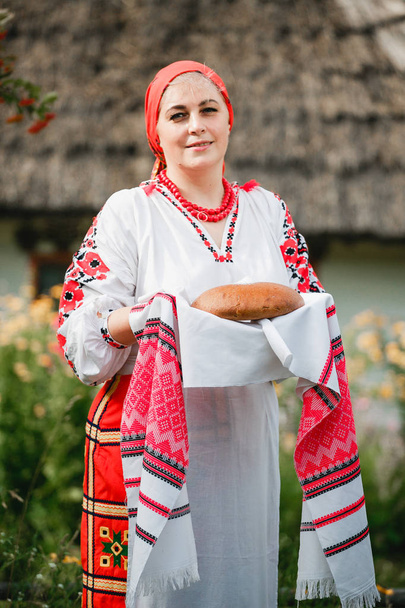 eine ukrainische frau in traditioneller tracht hält rushnyk mit brot und salz auf dem hintergrund eines alten hauses und blumen. - Foto, Bild