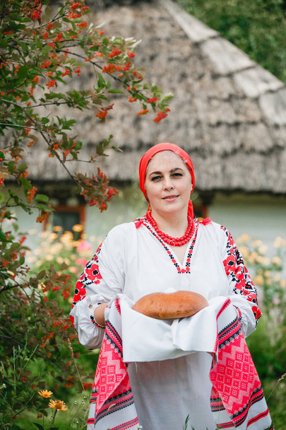 eine ukrainische frau in traditioneller tracht hält rushnyk mit brot und salz auf dem hintergrund eines alten hauses und blumen. - Foto, Bild