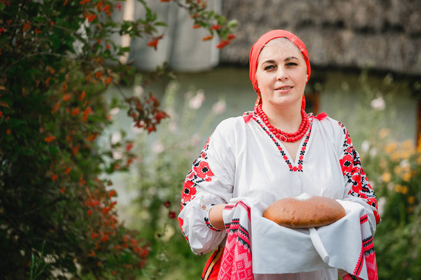 Украинка в традиционной одежде держит рушника с хлебом и солью на фоне старинного дома и цветов.
 - Фото, изображение