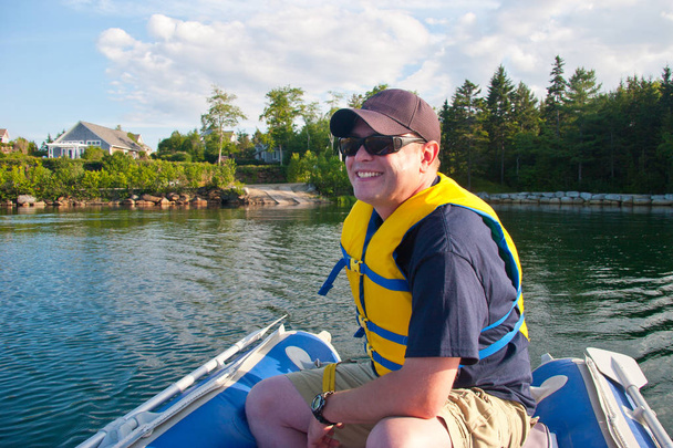 Egy ember, egy kalap és a mentőmellény viselése vigyorog, vidámság, vezet egy kis kék csónak körül egy tó vagy tenger.  - Fotó, kép