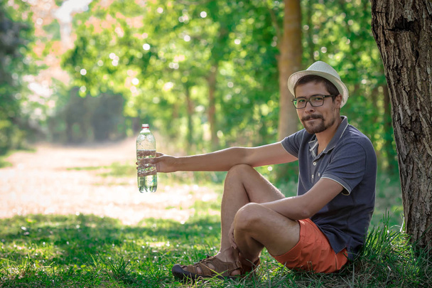 Usmívající se muž sedí v lese při pohledu na fotoaparát. chlap s vodou. poblíž strom a batoh - Fotografie, Obrázek