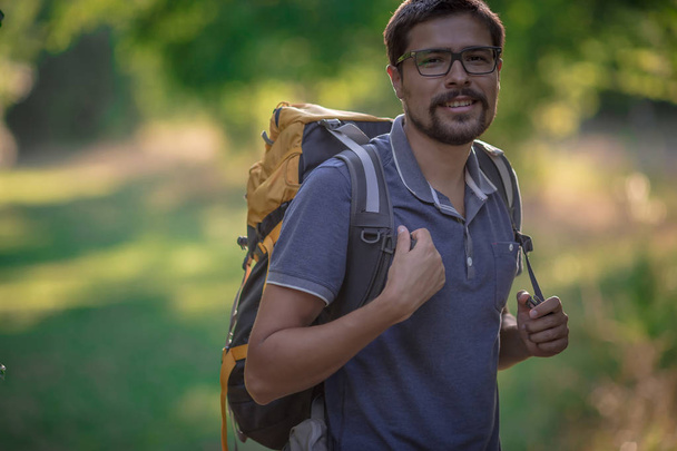Mladý usměvavý batoh muž v letní lesní přírodě. Šťastný pohledný muž dospělý student při pohledu na kameru chůze v lese pozadí. Školní taška nebo batohy cestovní koncept. - Fotografie, Obrázek