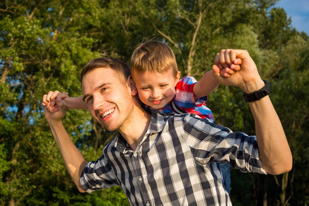 Щасливий молодий батько тримає свого сина на плечах і дивиться вгору. Маленький хлопчик сидить на плечах свого батька, імітуючи політ
 - Фото, зображення