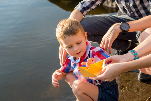 Dad and Mom give his son a paper boat to bring him down to the water. Mom, dad and their little son launch paper boats in a river in the summer - 写真・画像