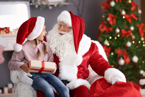 Little girl with gift box sitting on authentic Santa Claus' lap indoors - Photo, image