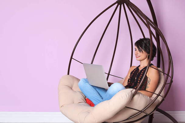 Young woman with modern laptop sitting in hanging chair near color wall - Foto, afbeelding