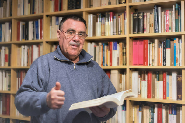 Portrait of senior man reading book in the hand on bookshelf background - Photo, Image