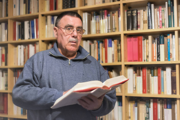 Porträt eines älteren Mannes, der Buch in der Hand auf Regalhintergrund liest - Foto, Bild