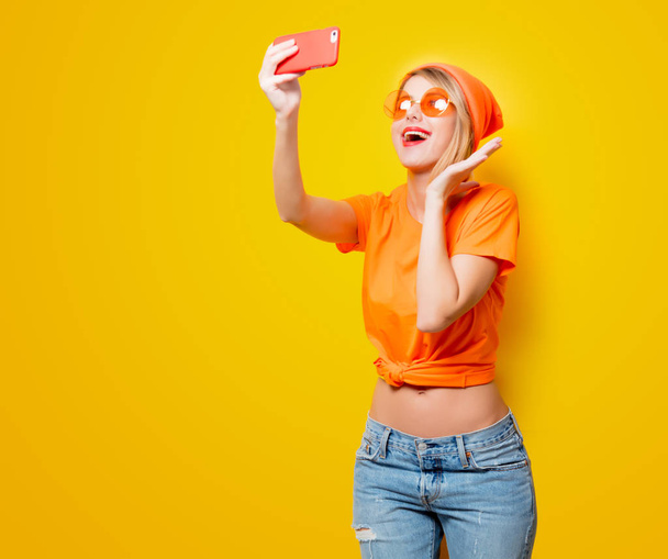 Young woman with orange glasses taking selfie with her phone on yellow background. Clothes in 1980s style - Foto, afbeelding