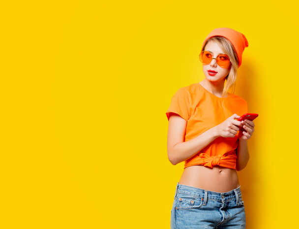 Mladá žena stylové oranžové brýle s jejím telefonem na žlutém podkladu. Oblečení ve stylu 1980s - Fotografie, Obrázek