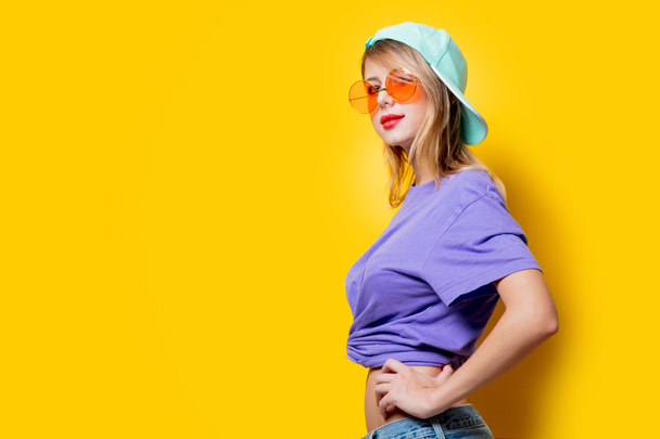 Young style girl with orange glasses and cap on yellow background. Clothes in 1980s style - Foto, Imagen