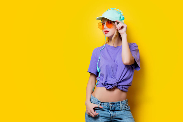 Stijlvolle jongedame met oranje bril en koptelefoon op gele achtergrond. Kleding in de stijl van de jaren 1980 - Foto, afbeelding