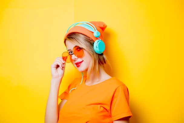 Jeune femme élégante avec des lunettes orange et des écouteurs sur fond jaune. Vêtements dans le style des années 1980
 - Photo, image