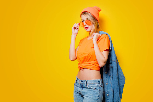 Mladá žena stylové oranžové brýle na žlutém podkladu. Oblečení ve stylu 1980s - Fotografie, Obrázek