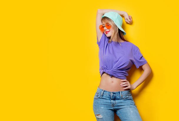 Jong meisje met oranje bril en pet op gele achtergrond. Kleding in jaren tachtig stijl - Foto, afbeelding
