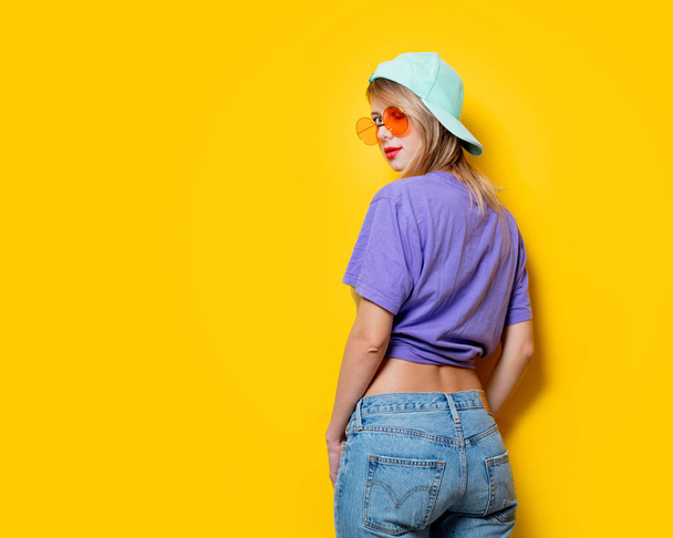 Sarı arka planda turuncu gözlüklü ve şapkalı genç bir kız. 1980 'lerin tarzında kıyafetler. - Fotoğraf, Görsel
