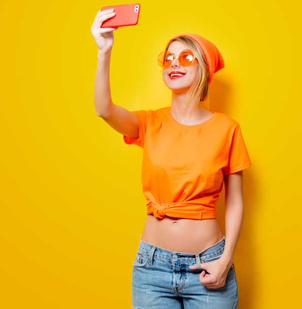 Молодая женщина в оранжевых очках делает селфи с телефоном на желтом фоне. Одежда в стиле 1980-х
 - Фото, изображение