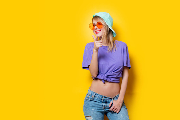 オレンジ色の眼鏡と黄色の背景にキャップを持つ若いスタイルの女の子。1980年代の服装 - 写真・画像