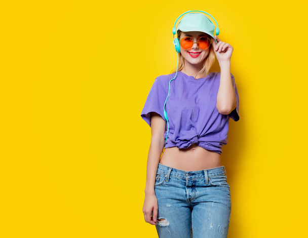 Young stylish woman with orange glasses and headphones on yellow background. Clothes in 1980s style - Фото, зображення