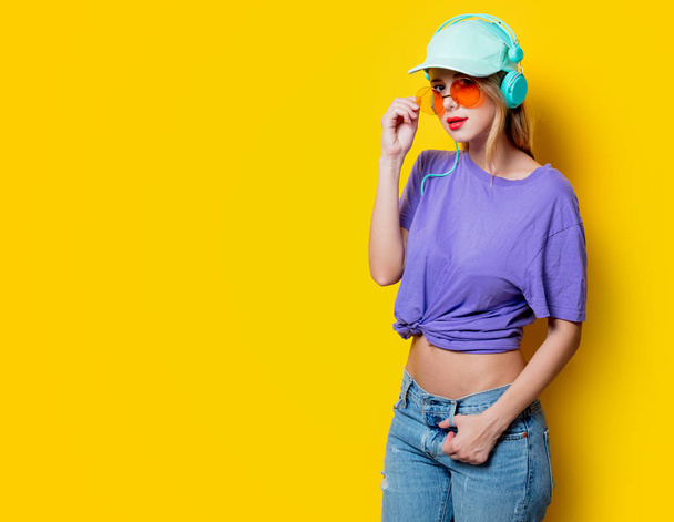 Mladá žena stylové oranžové brýle a sluchátka na žlutém pozadí. Oblečení ve stylu 1980s - Fotografie, Obrázek