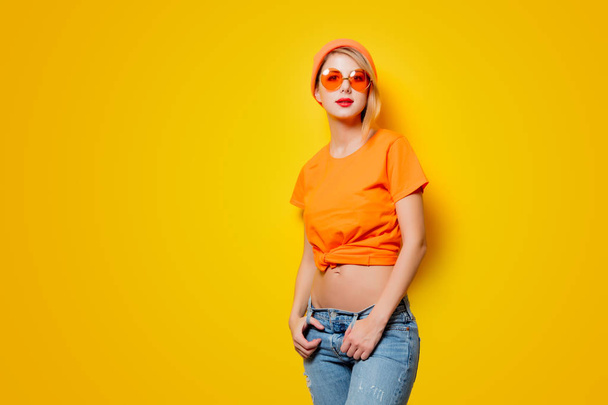Mujer joven y elegante con gafas de color naranja sobre fondo amarillo. Ropa en estilo de los años 80
 - Foto, imagen