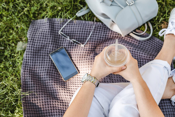 woman holding cup with cool drink while sitting on blanket on grass - Photo, Image