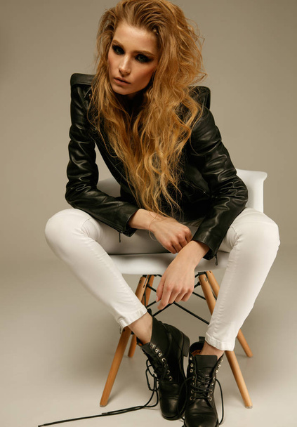 Female fashion model wearing grunge style leather jacket, white jeans and boots posing sitting on chair - Foto, Imagem