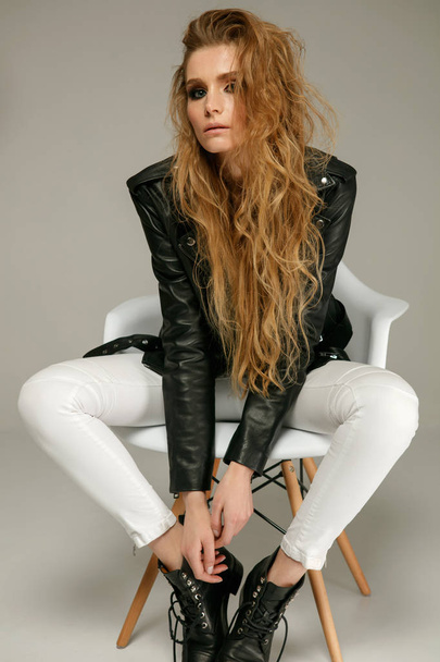 Female fashion model wearing grunge style leather jacket, white jeans and boots posing sitting on chair - Photo, image