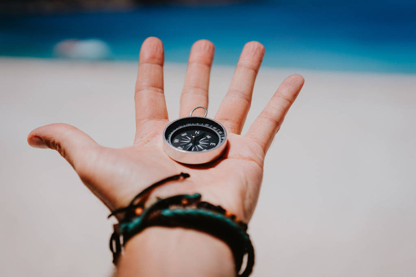Open palm with stretched fingers holding black metal compass against white sandy beach. Find your way or goal concept. Point of view pov - Фото, изображение
