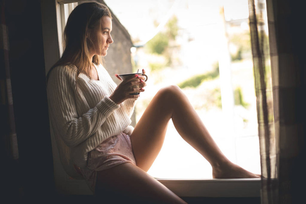 Donna premurosa che prende un caffè vicino alla finestra a casa
 - Foto, immagini