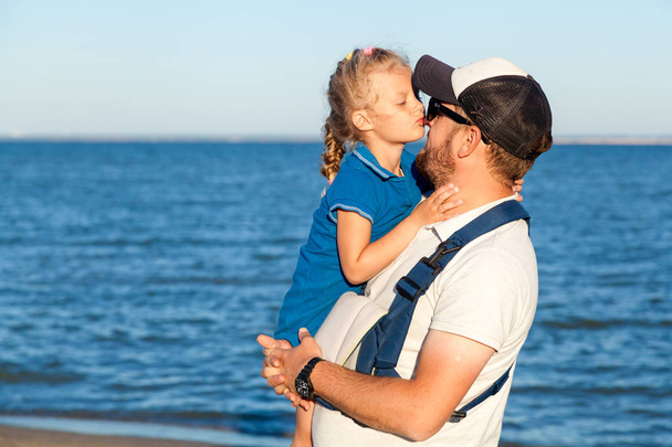 O jovem pai segura sua filha em seus braços e a beija contra o céu azul e o mar em um dia quente de verão.Conceito de férias de verão no mar e estilo ao vivo
 - Foto, Imagem