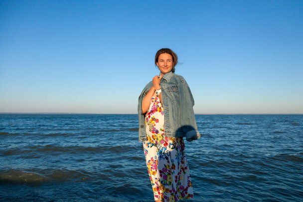 Een vrolijke dark-haired vrouw in de gekleurde wol en denim jasje glimlacht, wandelingen langs het strand en geniet van de stralende zon op een zomerdag. Concept van zomervakantie op zee en levende stijl - Foto, afbeelding
