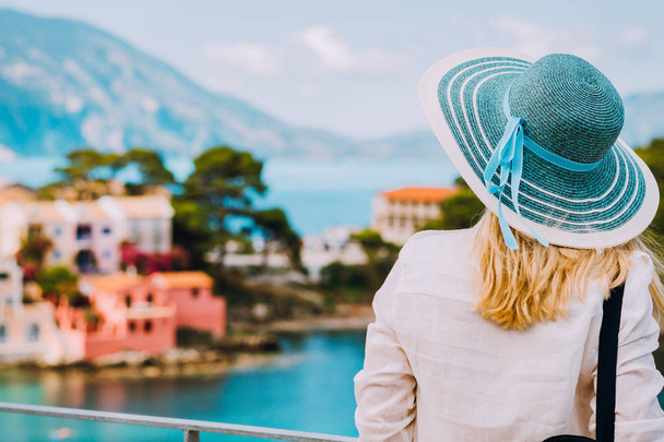 Tourist woman wear blue sunhat and white clothes admire view of colorful tranquil village Assos on sunny day. Stylish female visiting Kefalonia enjoying summer time on Greece travel vacation - Foto, Bild