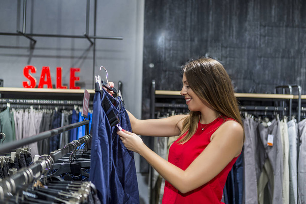 Attractive woman choosing cloths in shop. Beautiful woman checking tag in a boutique. Woman shopping trendy clothes. Shopaholic woman shopping clothes on sale. Woman in clothing store - Φωτογραφία, εικόνα