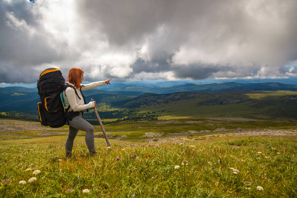 Piesze wycieczki kobieta z plecaka, ciesząc się, trekking i wskazujące kierunek strony Trek, w tle zielone pola, góry. Podróż życia i przetrwania koncepcja lusterka.  - Zdjęcie, obraz