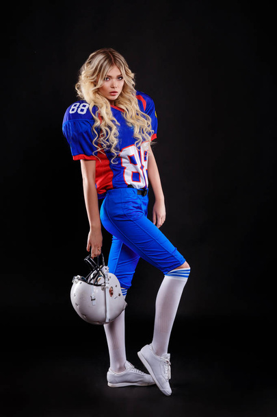 спортивная блондинка, изображающая американского футболиста на черном фоне. Красивая молодая женщина в американском футболе держит мяч
.  - Фото, изображение