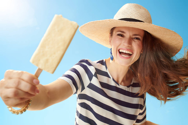happy young woman in straw hat against blue sky giving ice cream on a stick - 写真・画像