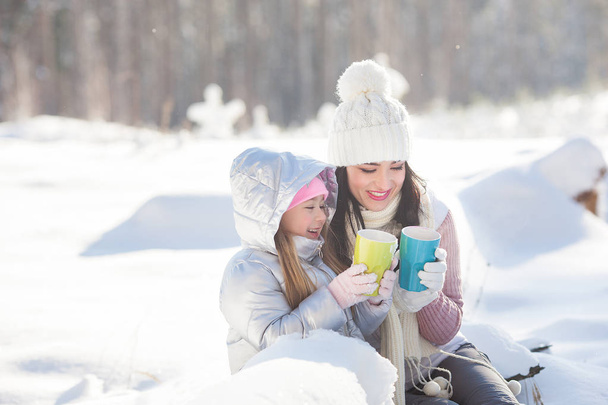 Mother and her daughter having fun outdoors in winter time. Pretty family drinking tea and cocoa - Zdjęcie, obraz