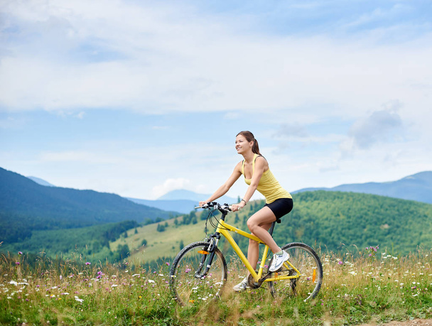 attraktive, glückliche Radfahrerin, die auf einem gelben Mountainbike auf einem grasbewachsenen Hügel fährt und den Sommertag in den Bergen genießt. Sport im Freien, Lifestylekonzept - Foto, Bild