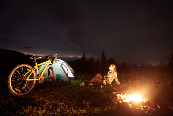 Jeune femme cycliste se reposant la nuit campant près d'un feu de camp allumé, tente touristique illuminée, VTT sous un beau ciel nocturne plein d'étoiles. Activité de plein air et concept touristique
 - Photo, image