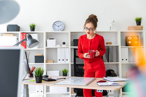 Egy fiatal lány áll közel a táblázat az irodájába, tart egy jelölőt és egy piros pohár a kezében, és úgy néz ki a mágnesfal. - Fotó, kép