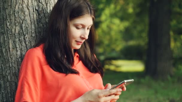 Beautiful young lady is using smartphone resting in park under tree and enjoying modern technology and summer nature. People, summertime and communication concept. - Video