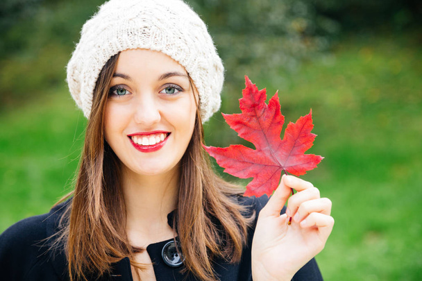 Beautiful young woman with woolen cap holding a red autumn leaf while smiling against a green background. Fresh skin and healthy smile. - Valokuva, kuva